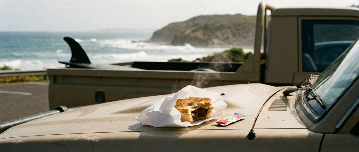 A half-eaten Australian meat pie steaming in its paper bag on the bonnet of a ute, surfboard in the tray and a point break behind