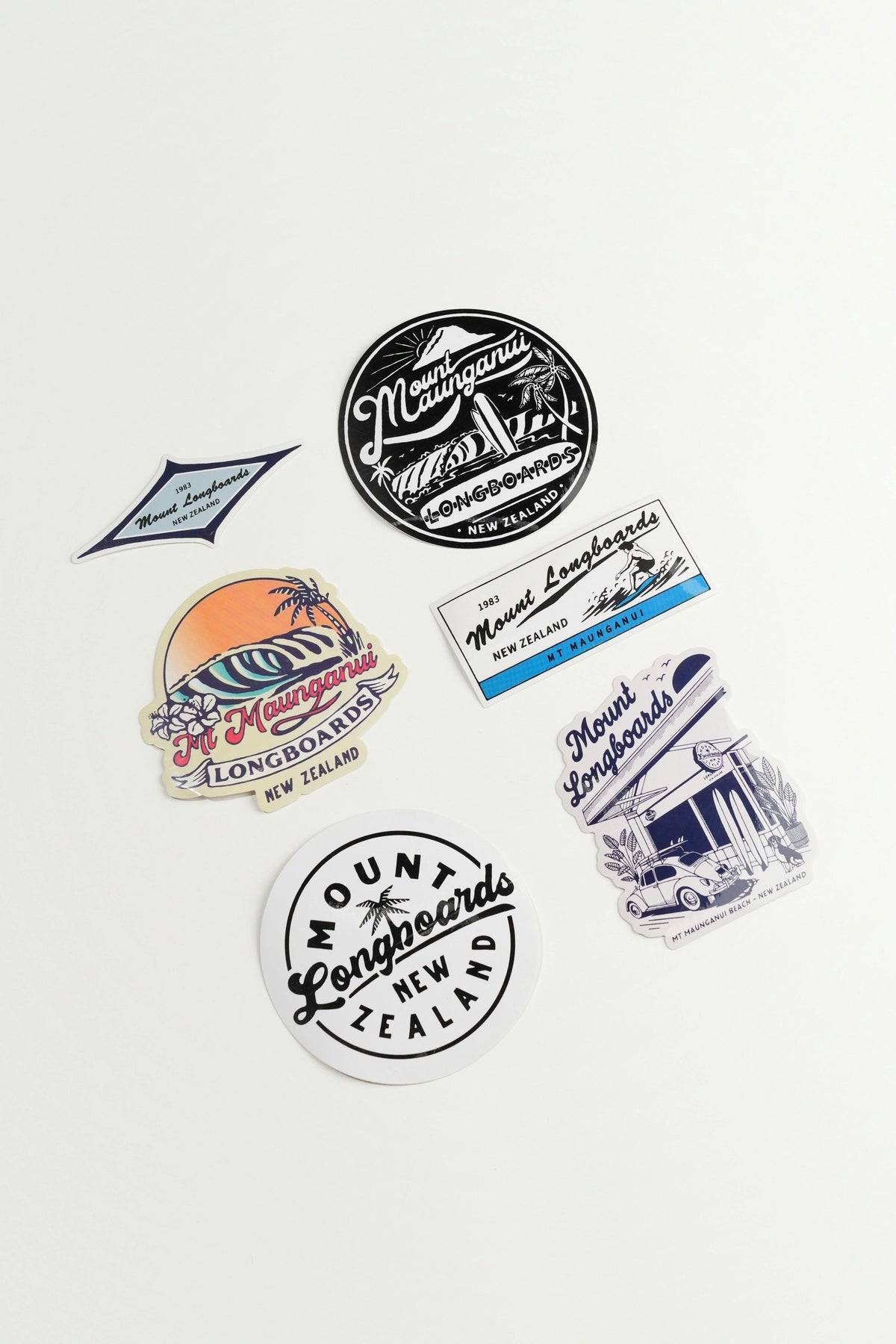 Six Mount Longboards stickers in various designs and colors, including black and white circular logos, a blue diamond sticker, a colorful sunset beach sticker, and a blue-and-white building illustration sticker, arranged on a clean white background.